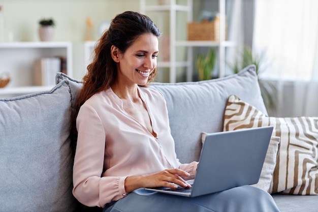 Lächelnde Frau, die zu Hause auf dem Sofa sitzt und einen Laptop benutzt