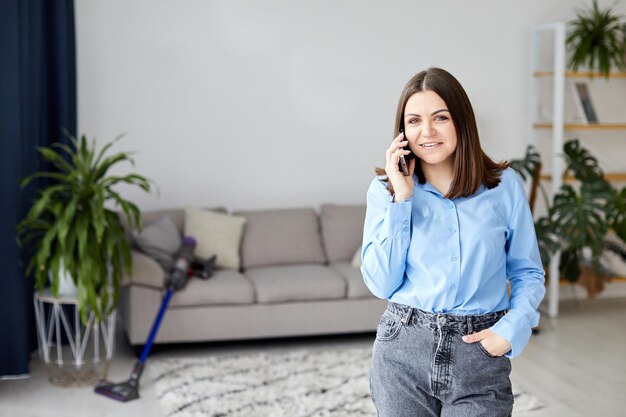 Lächelnde Frau, die zu Hause in der Nähe des Sofas telefoniert Weiblicher Klatsch auf dem Smartphone