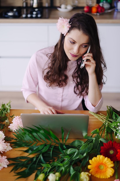 Lächelnde frau floristin inhaberin eines kleinen blumengeschäfts sie nimmt mit ihrem telefon und laptop bestellungen für ihren online-shop entgegen. arbeiten sie von zu hause aus