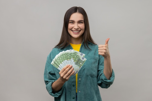 Lächelnde Frau hält viele Euro-Banknoten in der Hand und zeigt zufrieden den Daumen in die Kamera, weil sie im Lotto gewonnen hat