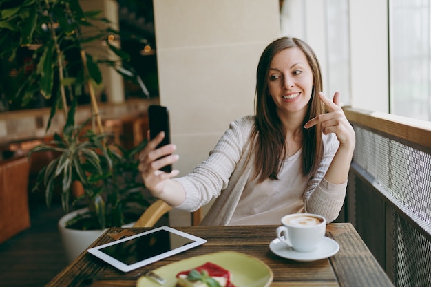 Lächelnde Frau im Café mit Tasse Cappuccino, Kuchen, Selfie auf dem Handy, Entspannung im Restaurant in der Freizeit. Weibliches Sitzen mit PC-Tablet-Computerrest im Café. Lifestyle-Konzept