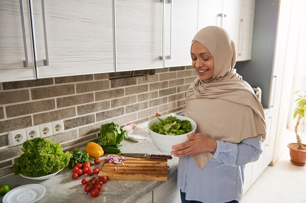 Lächelnde Frau im Hijab, die gegen ihre Hausküche steht und eine weiße Schüssel voll des gesunden veganen Salats hält