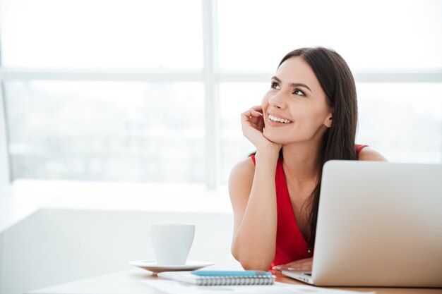 Lächelnde Frau im roten Hemd, die mit Laptop im Büro am Tisch sitzt und wegschaut