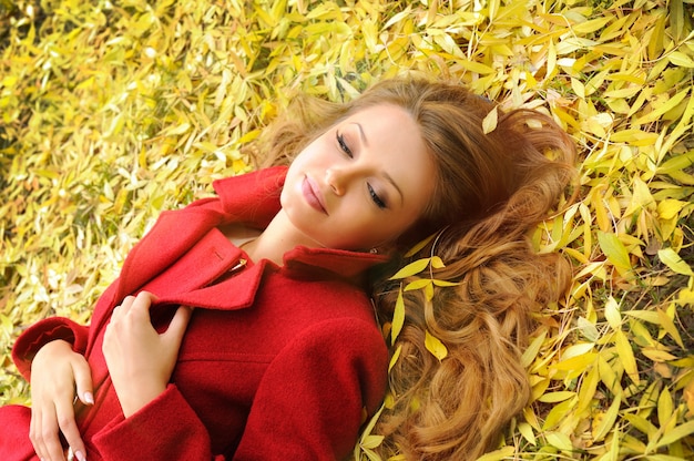 Lächelnde Frau im roten Mantel, der im Herbstlaub im Park liegt.