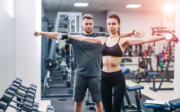 Lächelnde Frau im schwarzen Oberteil und in den Leggings, die Hanteln vor Trainer im Fitnessstudio halten.