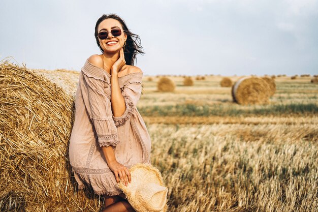 Lächelnde Frau in der Sonnenbrille mit nackten Schultern auf einem Hintergrund des Weizenfeldes und der Heuballen
