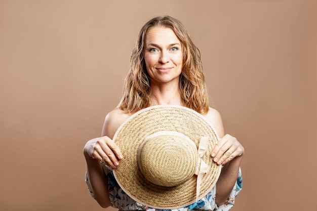 Lächelnde Frau in einem romantischen Sommerkleid schließt sich mit einem Strandstrohhut ab. Urlaub und Tourismus. Beigefarbener Hintergrund. Nahaufnahme