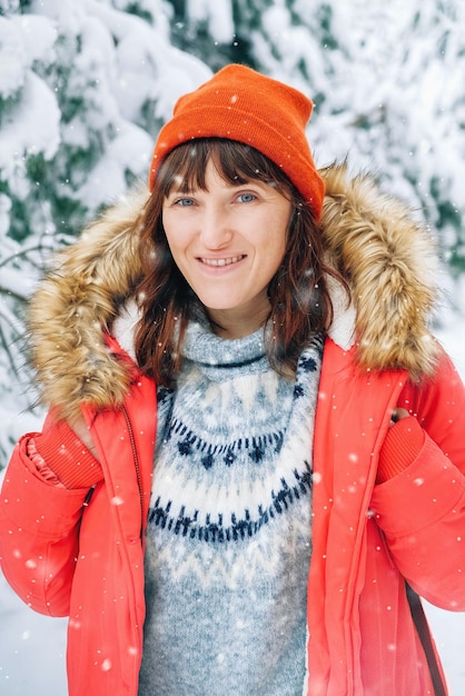 Lächelnde Frau in einer roten warmen Jacke und einem Hut auf einem Hintergrund des verschneiten Waldes