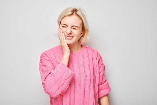 Lächelnde Frau in rosa Pullover mit der Hand auf der Wange auf einem grauen Hintergrund, die Zahnschmerzen ausdrückt