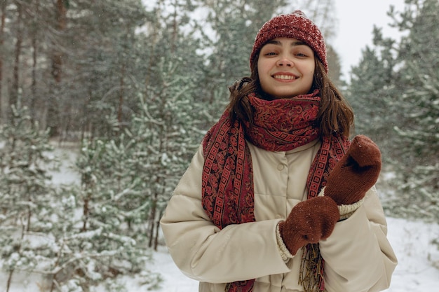 Lächelnde Frau in warmer Oberbekleidung und Fäustlingen, die im Winter im verschneiten Nadelwald stehen