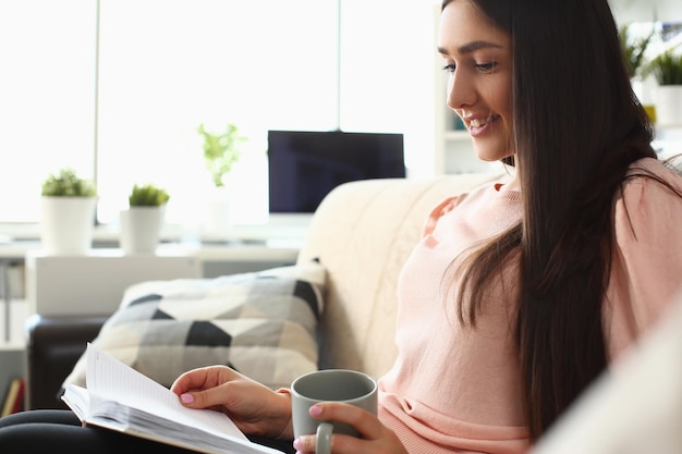 Lächelnde Frau liest ein Buch und hält eine Tasse Tee auf dem Sofa