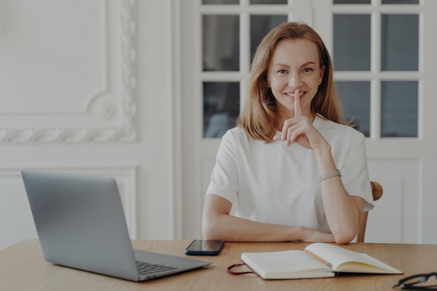 Lächelnde Frau macht Schweigegeste und bittet darum, geheim zu bleiben, wenn sie mit Laptop am Schreibtisch sitzt