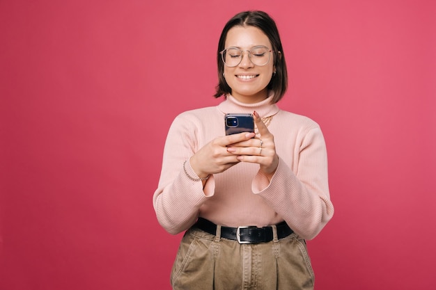 Lächelnde Frau mit Brille benutzt Telefon oder tippt eine Nachricht Studioaufnahme über rosa Hintergrund