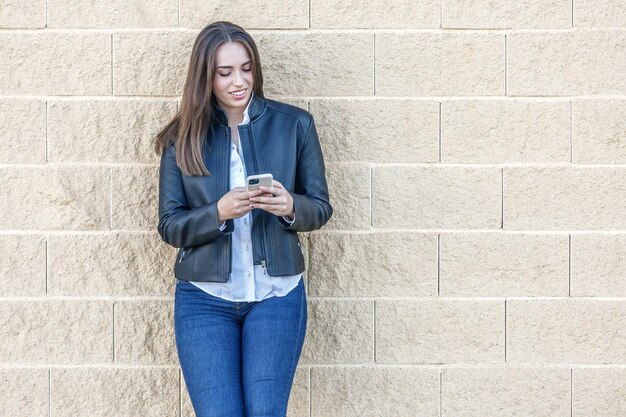 Lächelnde Frau mit einem Smartphone, die an einer Ziegelsteinmauer lehnt