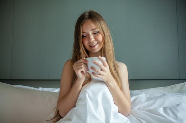 Lächelnde Frau mit einer Tasse heißem Kaffee in den Händen sitzt auf einem Hotelbett