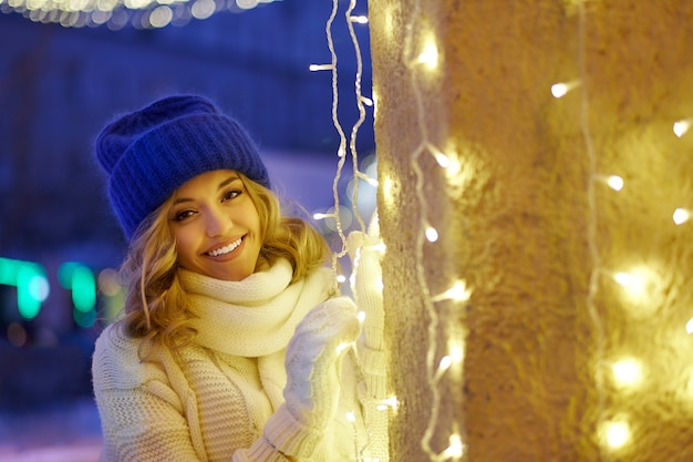 Lächelnde Frau mit Girlanden und Weihnachtsbeleuchtung auf festlicher Weihnachts- oder Neujahrsmesse. Dame mit klassischem, stilvollem Winterstrickpullover und -handschuhen