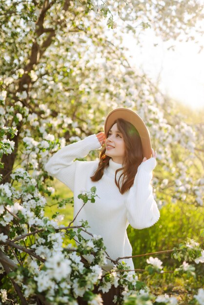 Lächelnde Frau mit Hut, die im blühenden Frühlingspark posiert Das Konzept der Entspannungsreise Modestil