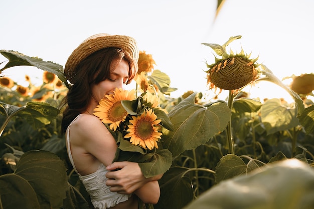 Lächelnde Frau mit Hut, die im Sonnenuntergang in einem Sonnenblumenfeld steht