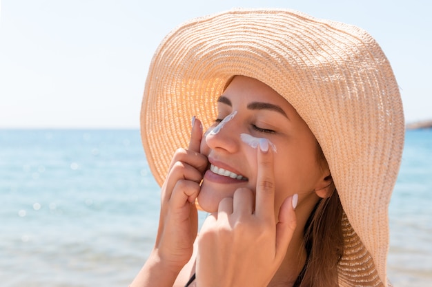 Lächelnde Frau mit Hut trägt Sonnenschutzmittel auf ihrem Gesicht auf. Indischer Stil.