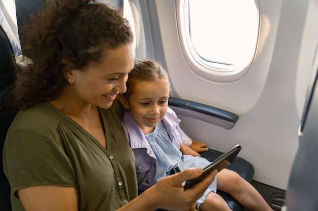 Lächelnde Frau mit ihrem Kind, die im Flugzeug sitzt und Smartphone benutzt