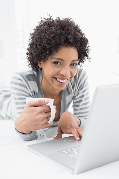 Lächelnde Frau mit Kaffeetasse unter Verwendung des Laptops im Bett