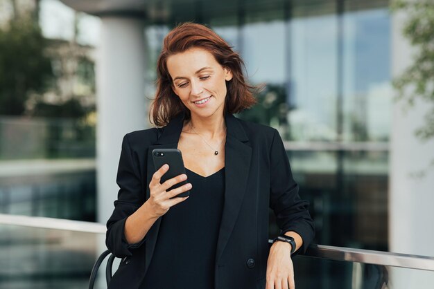 Lächelnde Frau mit roten Haaren, die formelle Kleidung trägt und auf ihr Smartphone schaut