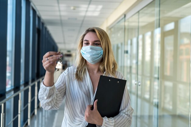 Lächelnde Frau mit Schutzmaske mit Hausschlüsseln im modernen Geschäftszentrum. Konzept von Immobilien und Büro