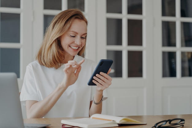 Lächelnde Frau mit Smartphone und mobilen Apps, die am Arbeitsplatz im Internet surfen