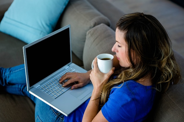 lächelnde Frau, sitzend auf ihrer Couch, nippend an Kaffee und schreibend