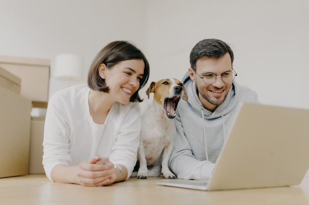 Lächelnde Frau und Mann arbeiten an moderner Laptop-Computer, Hund gähnt, kaufen Möbel für neue Wohnung, liegen auf dem Boden im geräumigen hellen Raum, haben frohe Ausdrücke. Einweihungsparty und Reparaturkonzept