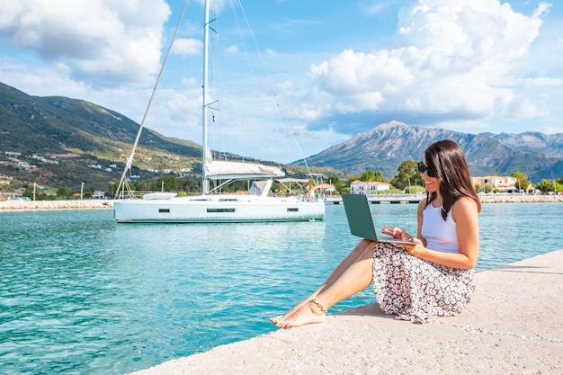 Lächelnde Freiberuflerin, die draußen am Laptop in den Bergen des Stadthafens und auf der Yacht im Hintergrund arbeitet