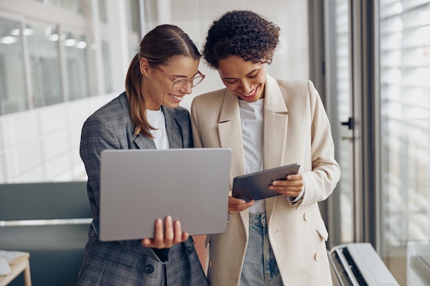 Lächelnde freiberufliche Mitarbeiter stehen im Coworking und arbeiten auf einem digitalen Tablet und Laptop