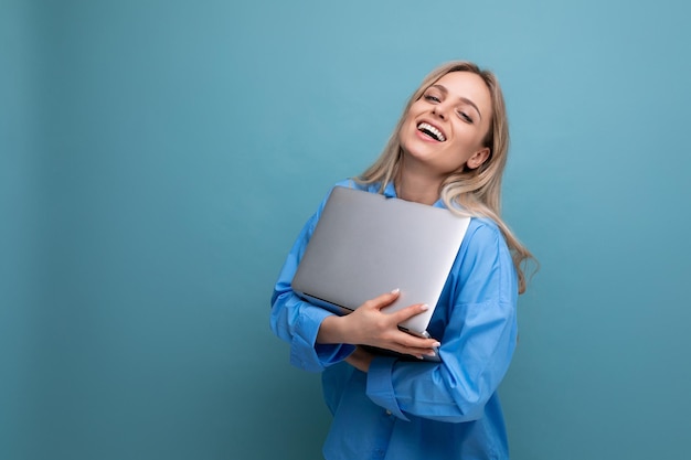 Lächelnde fröhliche blonde Studentin mit einem Laptop-Computer auf einem hellblauen Hintergrund