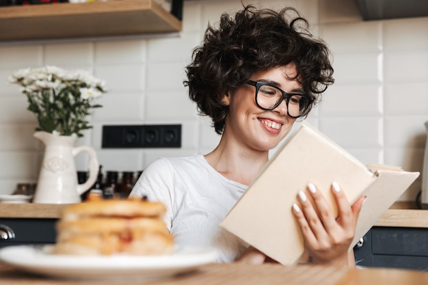 Lächelnde fröhliche Frau, die leckeres Frühstück beim Sitzen in der Küche zu Hause hat und ein Buch liest