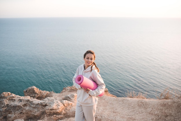 Lächelnde fröhliche Frau hält Yoga-Matte tragen Trainings-Hoodie posiert über Meer