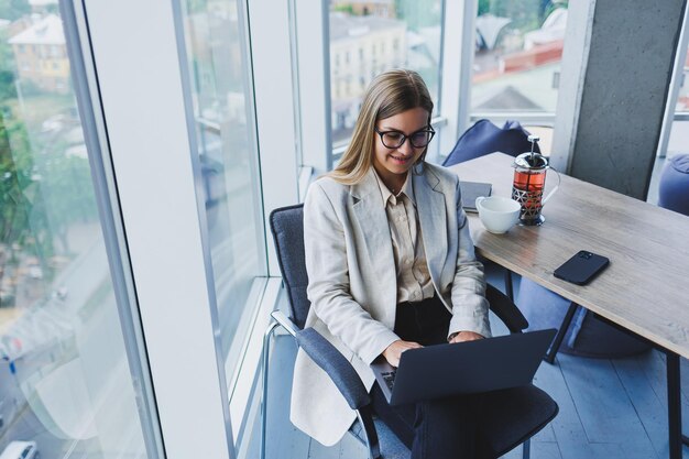 Lächelnde Geschäftsfrau, die Laptop während der Arbeit im Büro betrachtet Das Konzept einer modernen erfolgreichen Frau Die Idee des Geschäfts und des Lebens eines Unternehmers Junge Frau am Tisch mit offenem Laptop