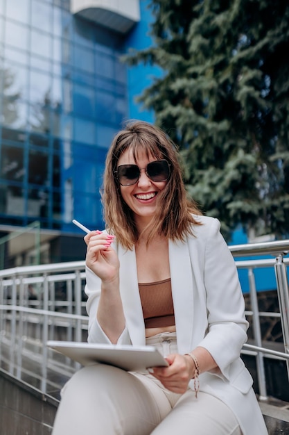 Lächelnde Geschäftsfrau in weißem Anzug und Sonnenbrille, die an einem digitalen Tablet in der Nähe eines modernen Bürogebäudes arbeitet