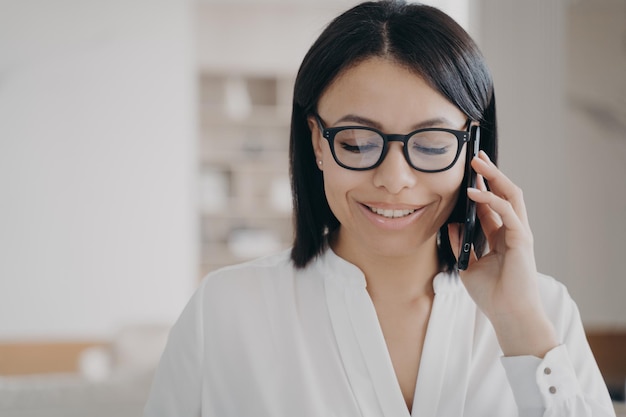 Lächelnde Geschäftsfrau mit Brille, die auf dem Smartphone mit dem Kunden spricht, der den Anruf im Büro entgegennimmt