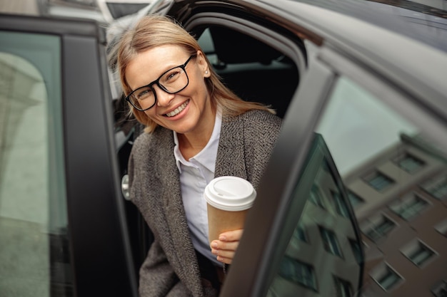 Lächelnde Geschäftsfrau mit Brille und Kaffee zum Mitnehmen, der aus dem Auto steigt