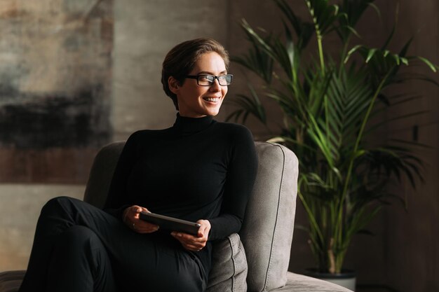 Lächelnde Geschäftsfrau schaut weg, während sie drinnen sitzt und ein digitales Tablet in der Hand hält