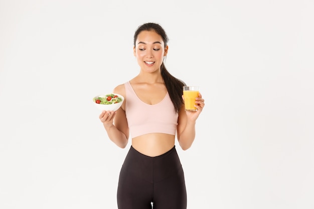 Lächelnde gesunde und schlanke brünette asiatische Mädchen wie Fitness, ins Fitnessstudio gehen und auf Diät sein, Salat mit Orangensaft halten, stehen