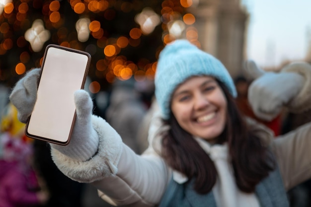 Lächelnde glückliche Frau an der Weihnachtsstadtmesse, die Telefon mit weißem Bildschirm hält