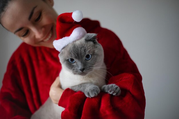 Lächelnde, glückliche Frau umarmt ihre süße schottische Kurzhaarkatze in Santa Christmas hat Spaß mit Haustier zu Hause Weihnachts- und Neujahrskonzept