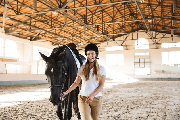 lächelnde glückliche junge blonde schöne Frau mit Hut mit Pferd in der Landschaft in Corral auf dem Trainingsgelände.