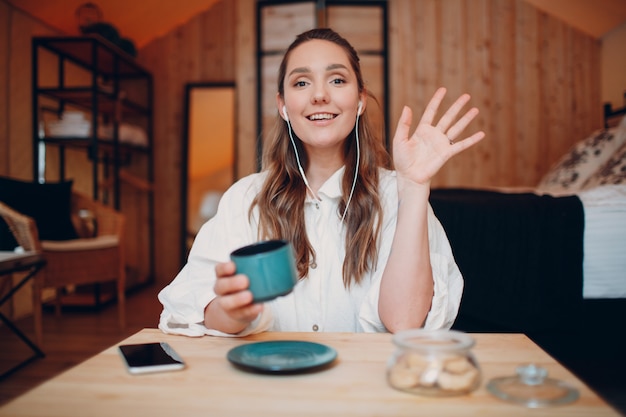 Lächelnde, glückliche junge Frau, die zu Hause hinter einem Computer-Laptop am Tisch sitzt und über Videoanrufmädchen mit einer Tasse Tee oder Kaffee spricht, die online vor der Webcam drinnen spricht