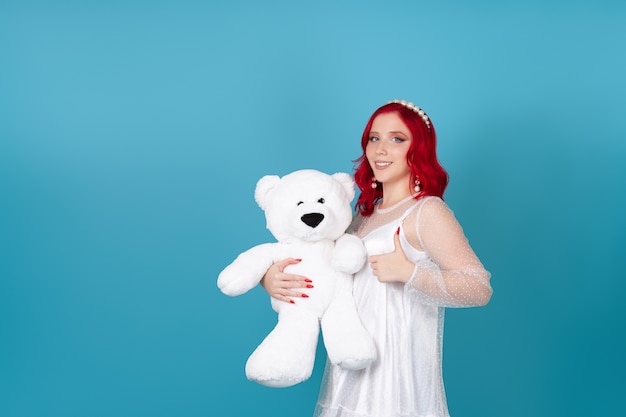 Lächelnde glückliche junge Frau im weißen Kleid mit den roten Haaren hält weißen Teddybär und gibt Daumen auf
