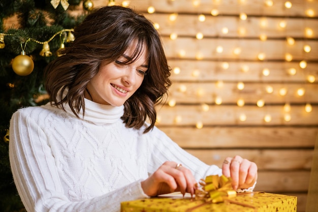 Lächelnde glückliche junge Frau im Winterpullover mit goldenem Geschenk, während sie zu Hause auf dem Boden sitzt...