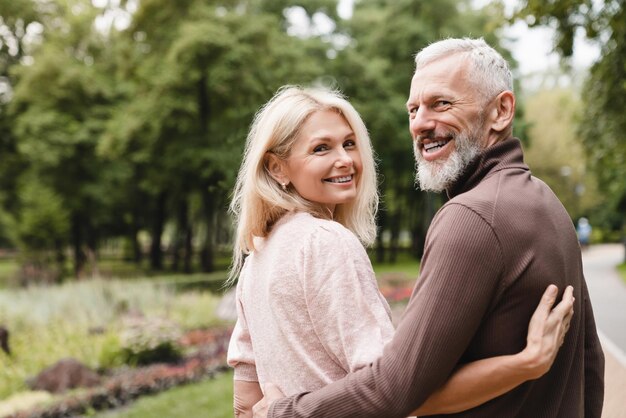 Lächelnde, glückliche, kaukasische, reife Ehepartner, die sich umarmen und umarmen, während sie zusammen auf einem Date im Park spazieren gehen, die Liebe und Beziehung verbinden.