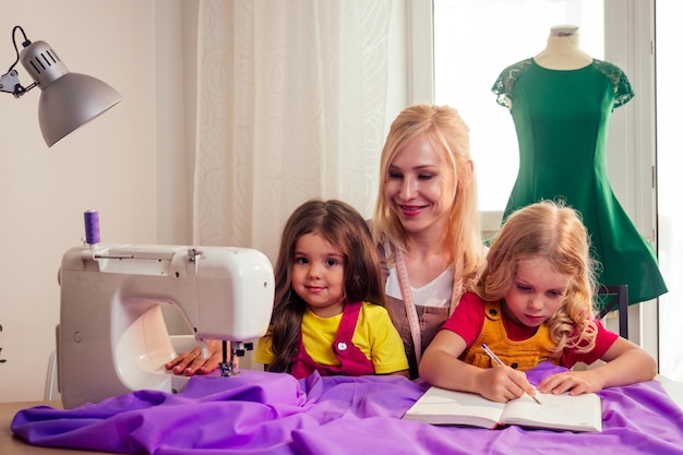 Lächelnde, glückliche, Multitasking-blonde Mutter bringt einer kleinen Schwester bei, selbst an einer Nähmaschine zu nähen