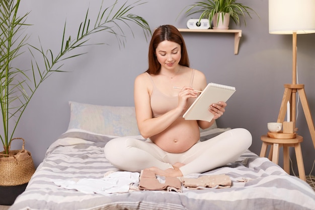 Lächelnde, glückliche, schwangere, braunhaarige, kaukasische Frau, die im Bett sitzt und ein Baby erwartet, das ein Notizbuch aus Papier hält, das sich auf die Geburt vorbereitet